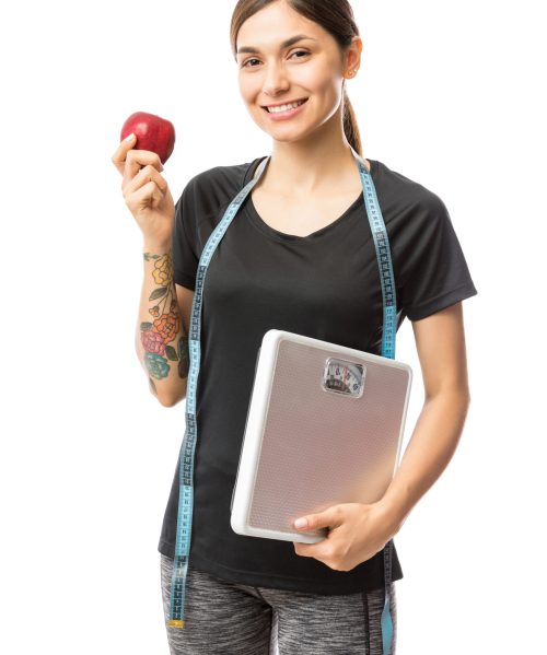 Portrait of smiling young fit woman holding apple and weight scale over white background